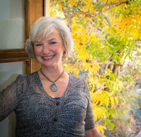 White-haired woman stands in front of open window with green and yellow flora behind her