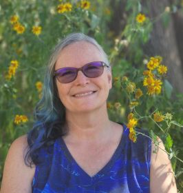 Headshot of Paige E. Ewing (dark blue tank top, shoulder-length grey hair, sunglasses) with a green leafy background.